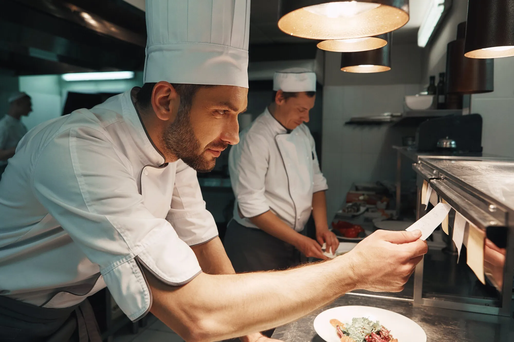 Koch prüft in einer vollen Restaurantküche einen Bon, hinter ihm arbeitet das Team