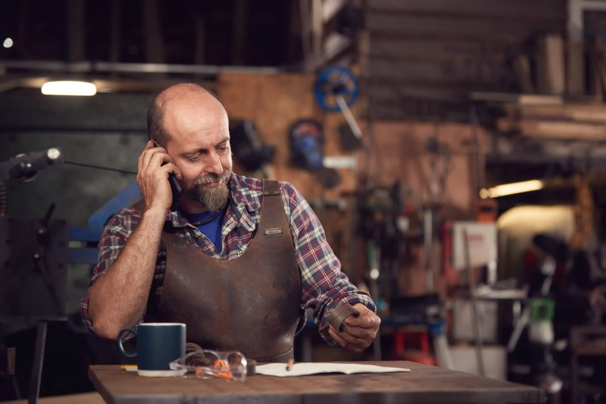 Schmied telefoniert am Werktisch und hält dabei ein Werkstück